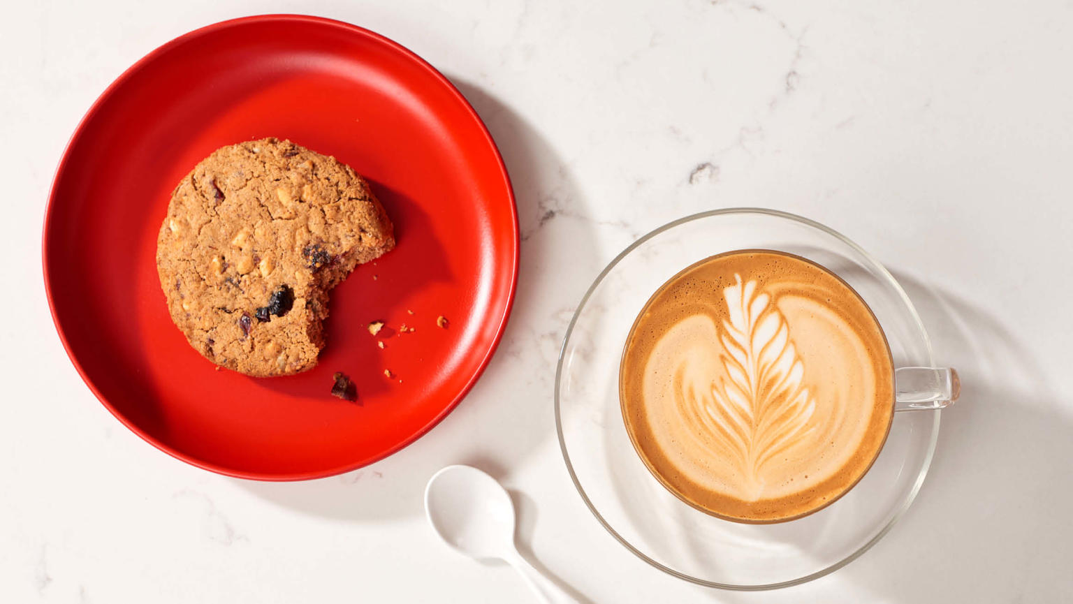 A beautifully crafted cup of coffee with intricate latte art, accompanied by a red plate showcasing a delectable cookie with a bite missing.