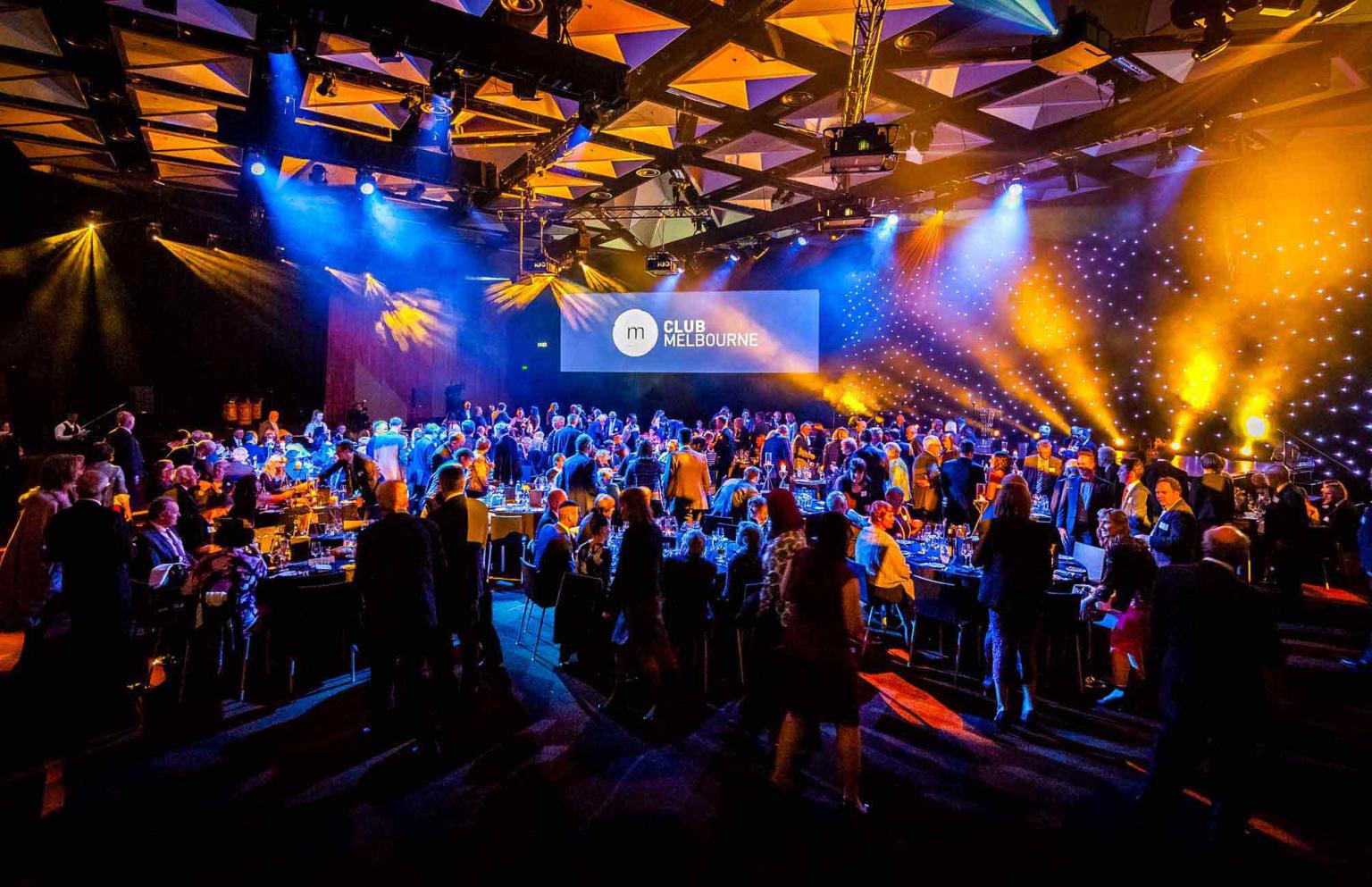 Large crowd seated at a formal gala dinner, with blue and yellow lights over head. In the background a screen displays the Club Melbourne logo. 