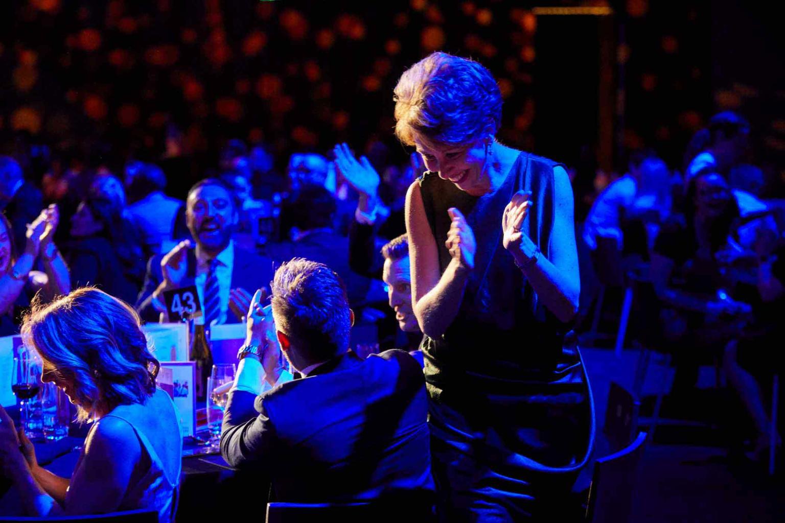 A woman stands clapping her hands looking down at man who is seated at a table. A large crowd sits behind at more formal dinner tables. There is blue lighting that colours the crowd. 