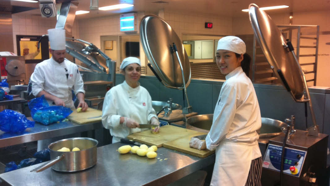 Three chefs in an industrial kitchen chopping potatoes and placing them in pots for cooking. 