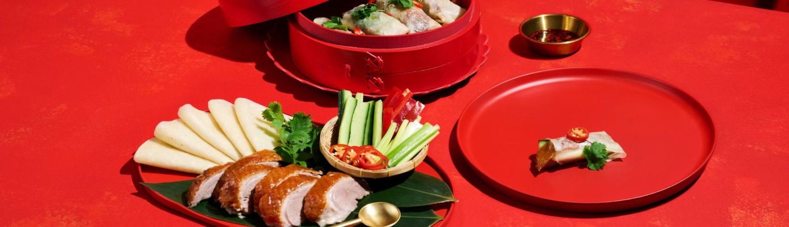 A red table is full of dishes. One is a sichuan roasted duck breast wrapped in a traditional pancake and another red bowl full of poached chicken rice paper roll with Vietnamese mint and nuoc cham dipping sauce.