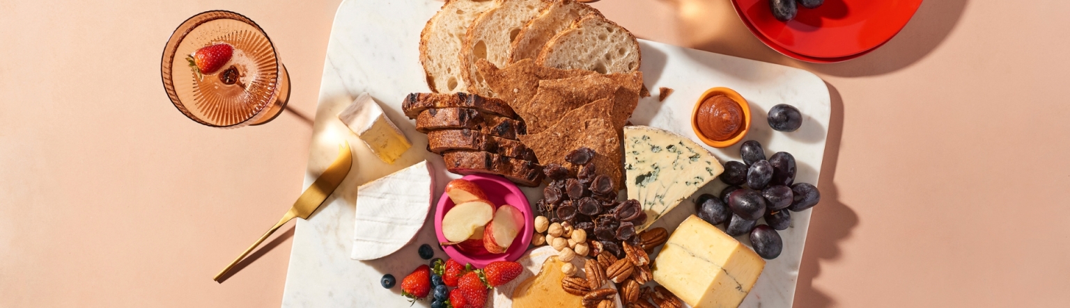 A showcase of cheese served with fruit, nuts and house-baked sourdough bread on a white marble platter.