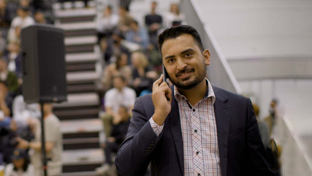A man in a shirt and blazer happily talks on the phone while looking towards the camera. In the background, a sizeable crowd is seated on levelled stands, facing large speakers positioned before them.