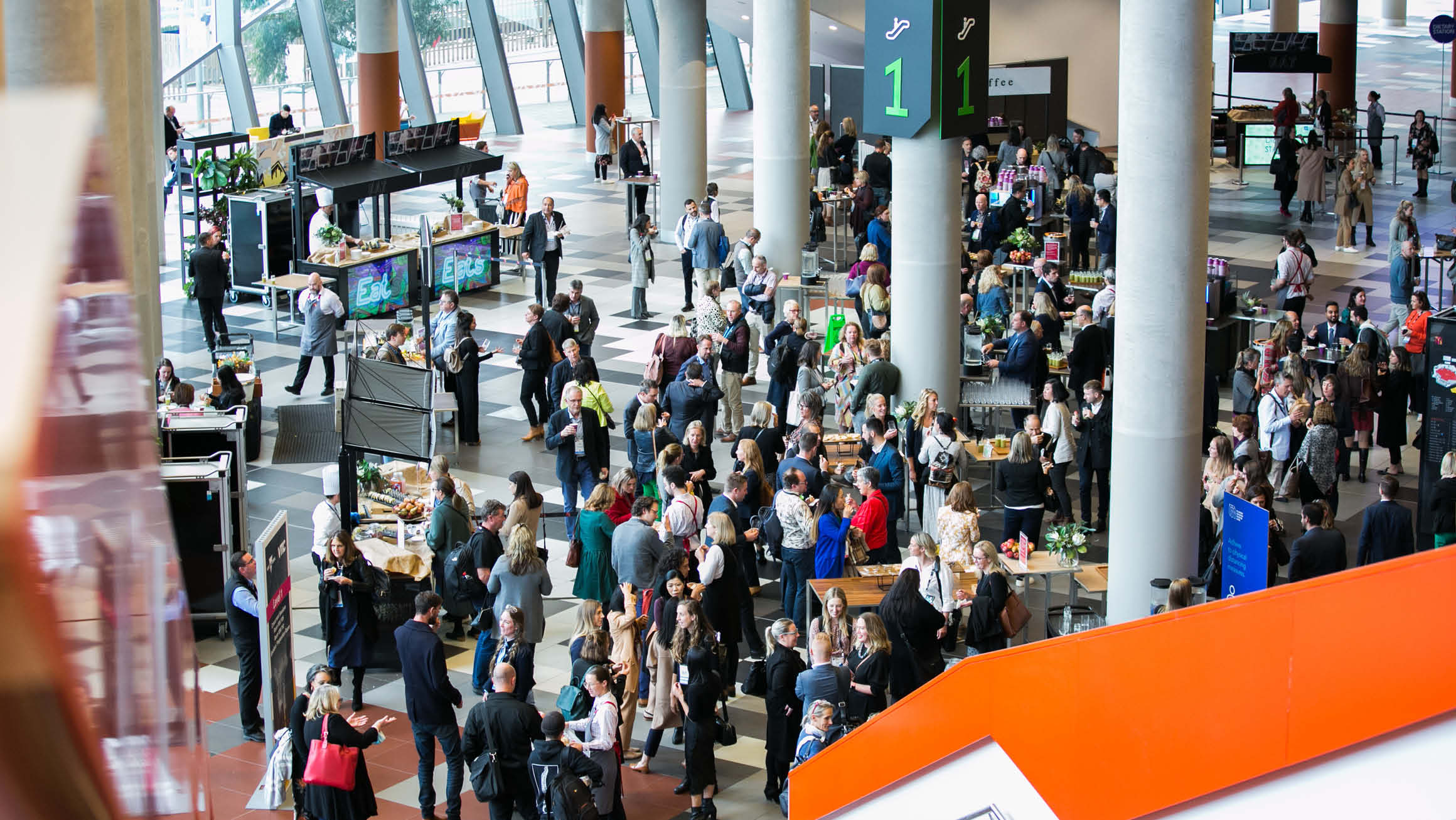 An overhead view capturing a spacious foyer bustling with a large crowd of people. Concrete pillars and food stations are strategically placed throughout the area, creating a dynamic and inviting atmosphere. The image offers a comprehensive perspective of the lively scene from above.