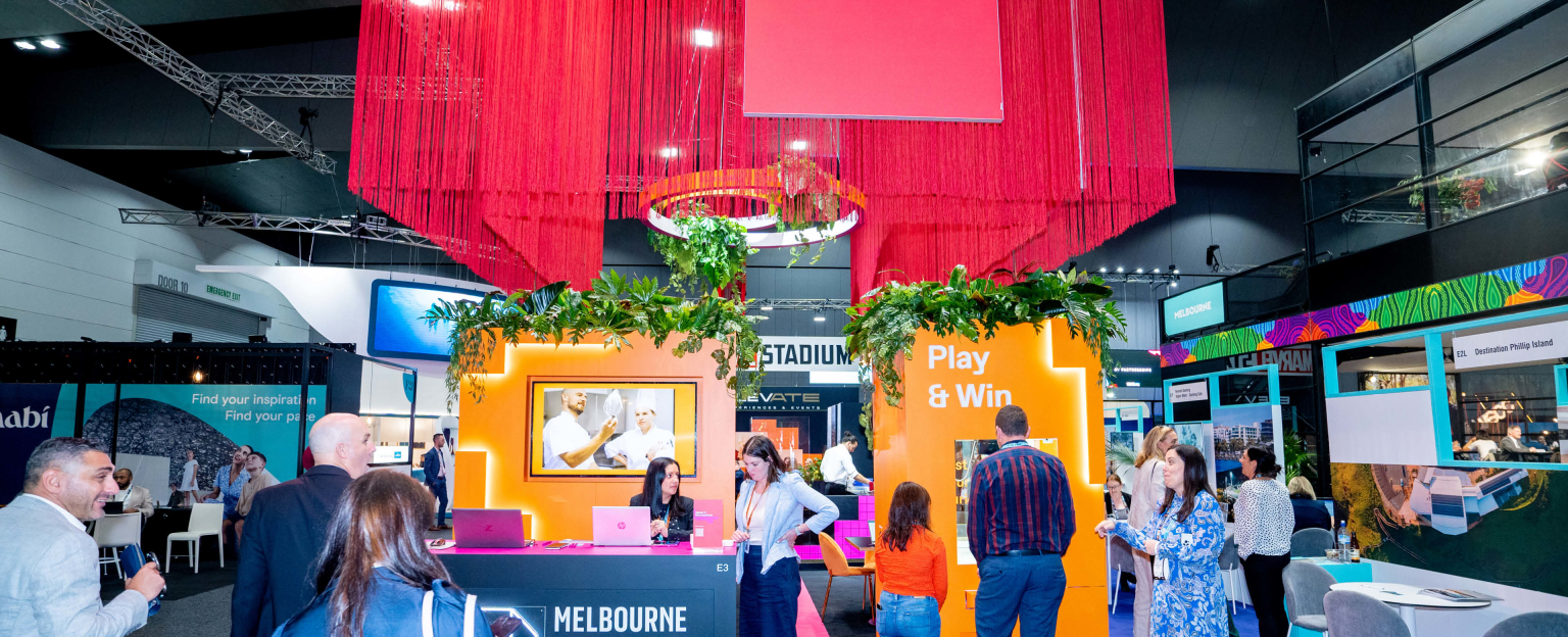 An eye-catching exhibition stand with a captivating arrangement: long red fringing suspended from the ceiling, an inviting orange wall featuring a walk-through cutout adorned with lush greenery, and a dynamic digital screen enhanced by neon lights. On the left side, a vibrant pink and black table proudly showcases a neon logo displaying 'Melbourne Convention and Exhibition Centre.' To the right, an enthralling claw drop game entices visitors with the enticing phrase 'Play & Win' displayed above it. Excited individuals surround the stand, contributing to the energetic atmosphere.