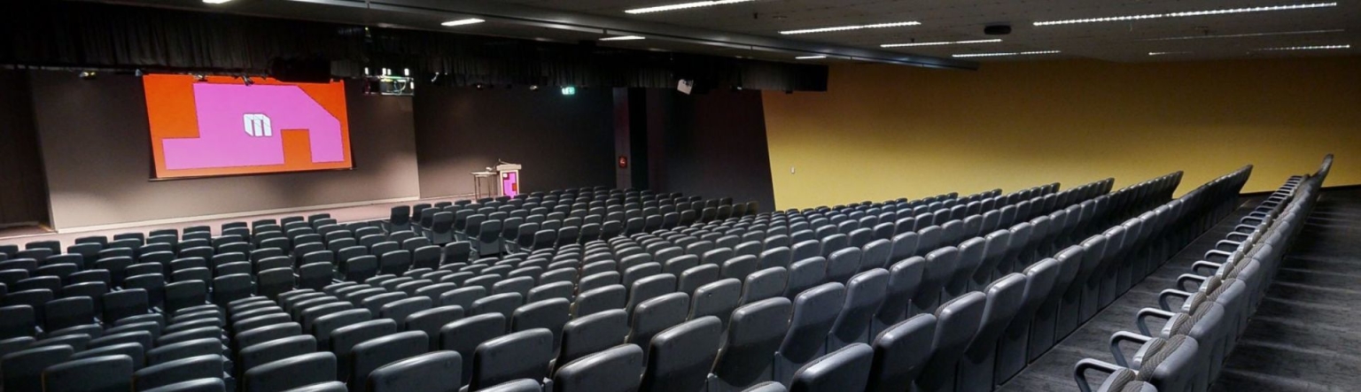 Auditorium room full of rows of black seats facing a large projector screen at the front of the room over a stage. The MCEC logo is displayed on the screen over a pink and red shape design. 