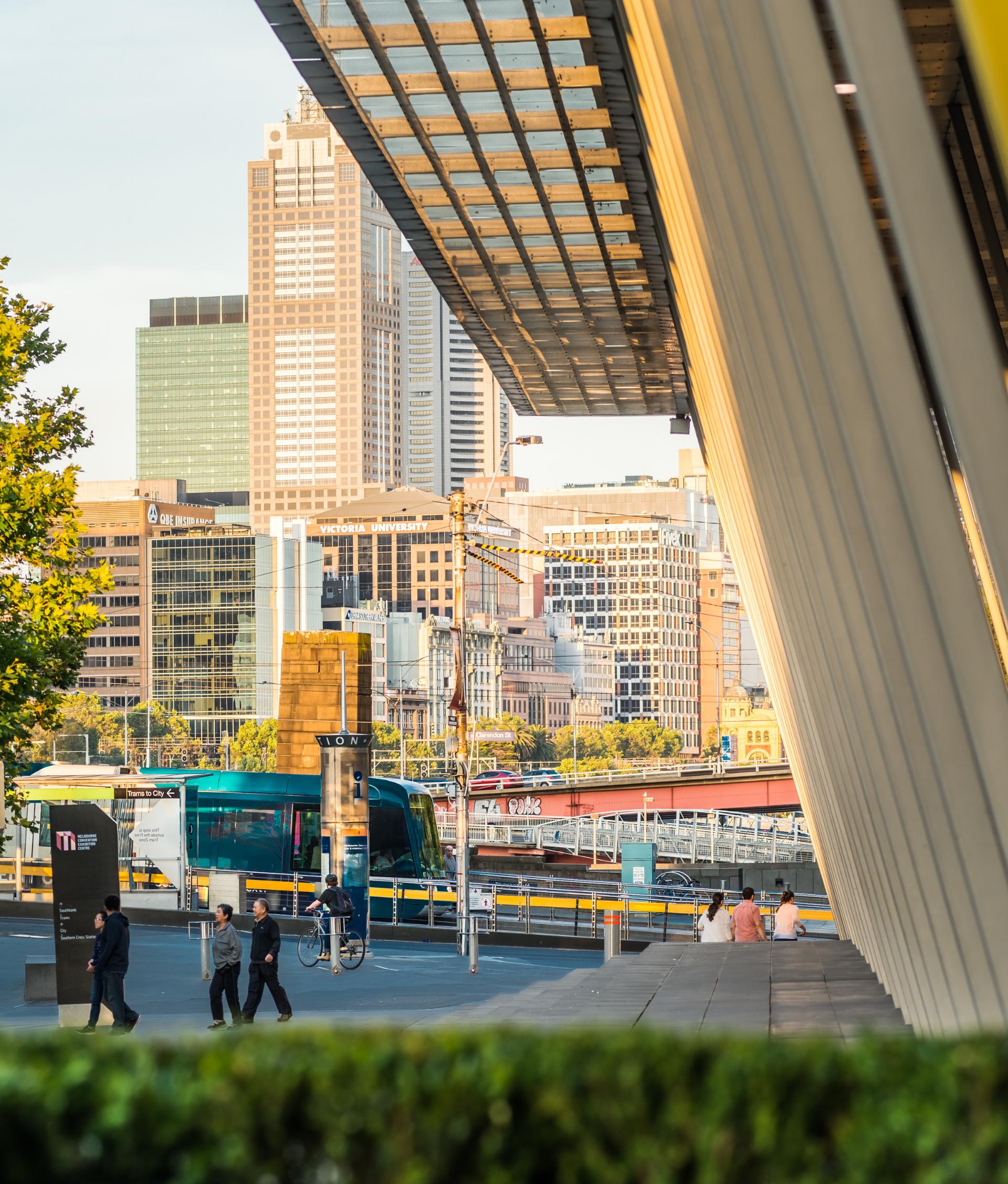 Outside view of the columns of the Exhibition Building, looking towards the tram stop near Crown. Tall buildings are in the distance and a green bush is blurry in the foreground. 