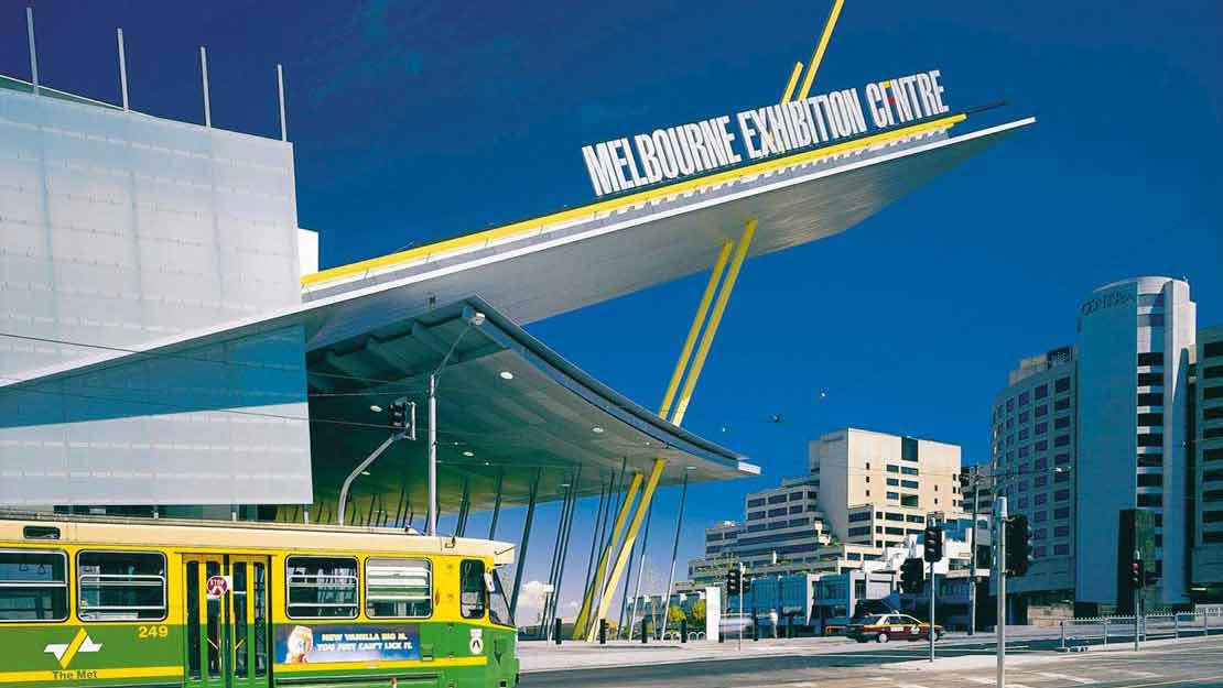 Tram is moving across the foreground outside the Melbourne Exhibition Building Clarendon Street entry. Blue skies are seen behind. 