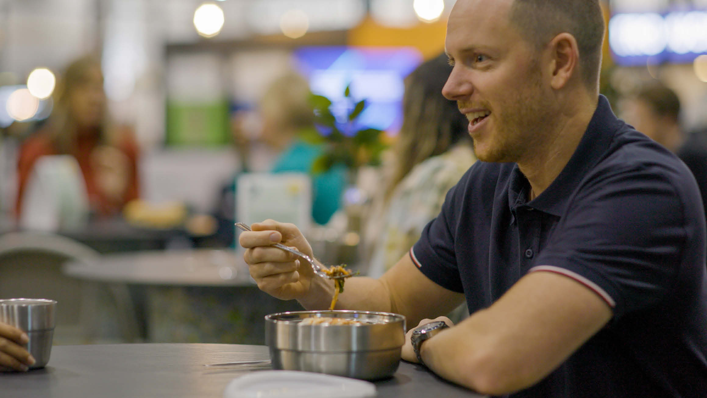 Man in polo shirt sitting at a table eating food from a stainless steal bowl smiling to someone off camera. 