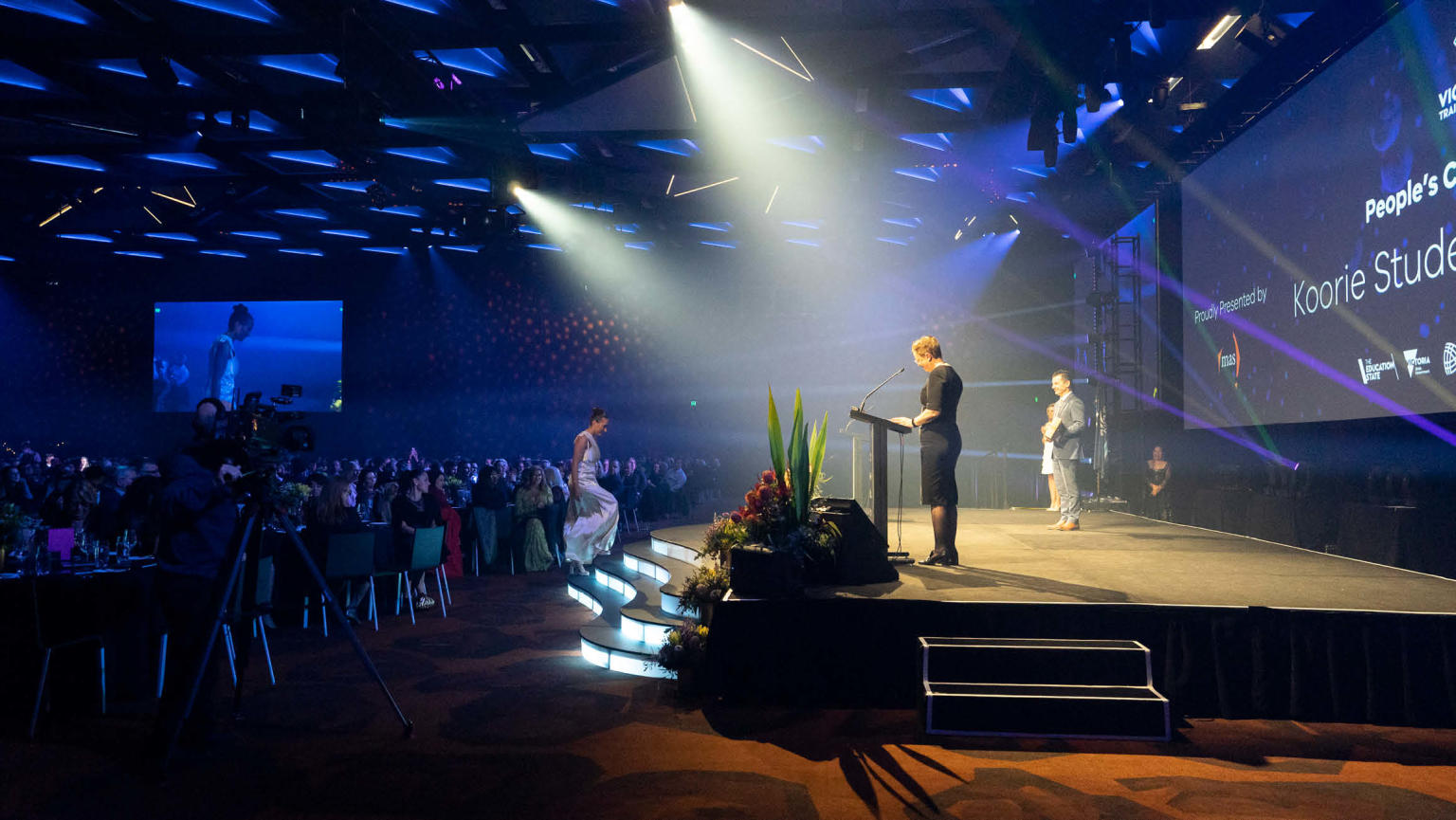 A focused spotlight illuminating a stage where a woman stands behind a lectern, addressing the audience through a microphone. The attentive audience is seated at round tables, engaged in listening.