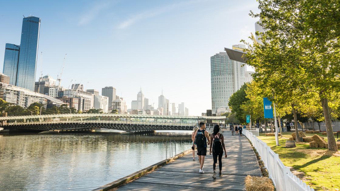 People walking along a boardwalk next to a river. City views are in the background and bright green grass and trees along the side. DFO flags wave on poles along the path. 