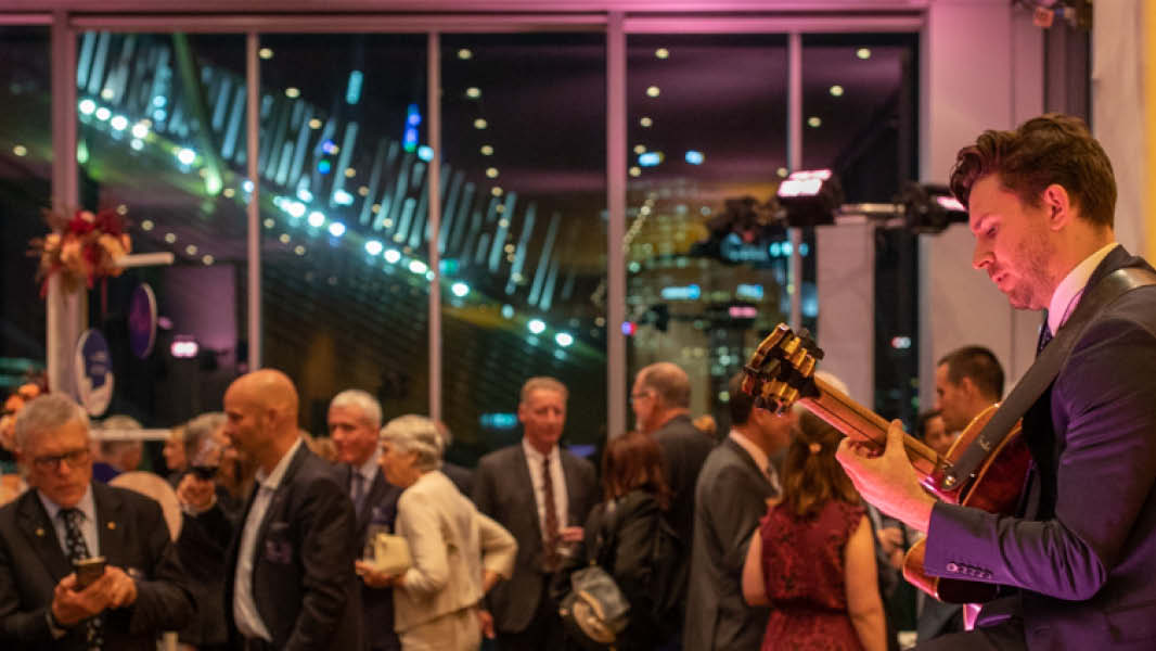 Musician playing guitar stands to the right of the image, performing to a crowd of people in formal attire socialising. 