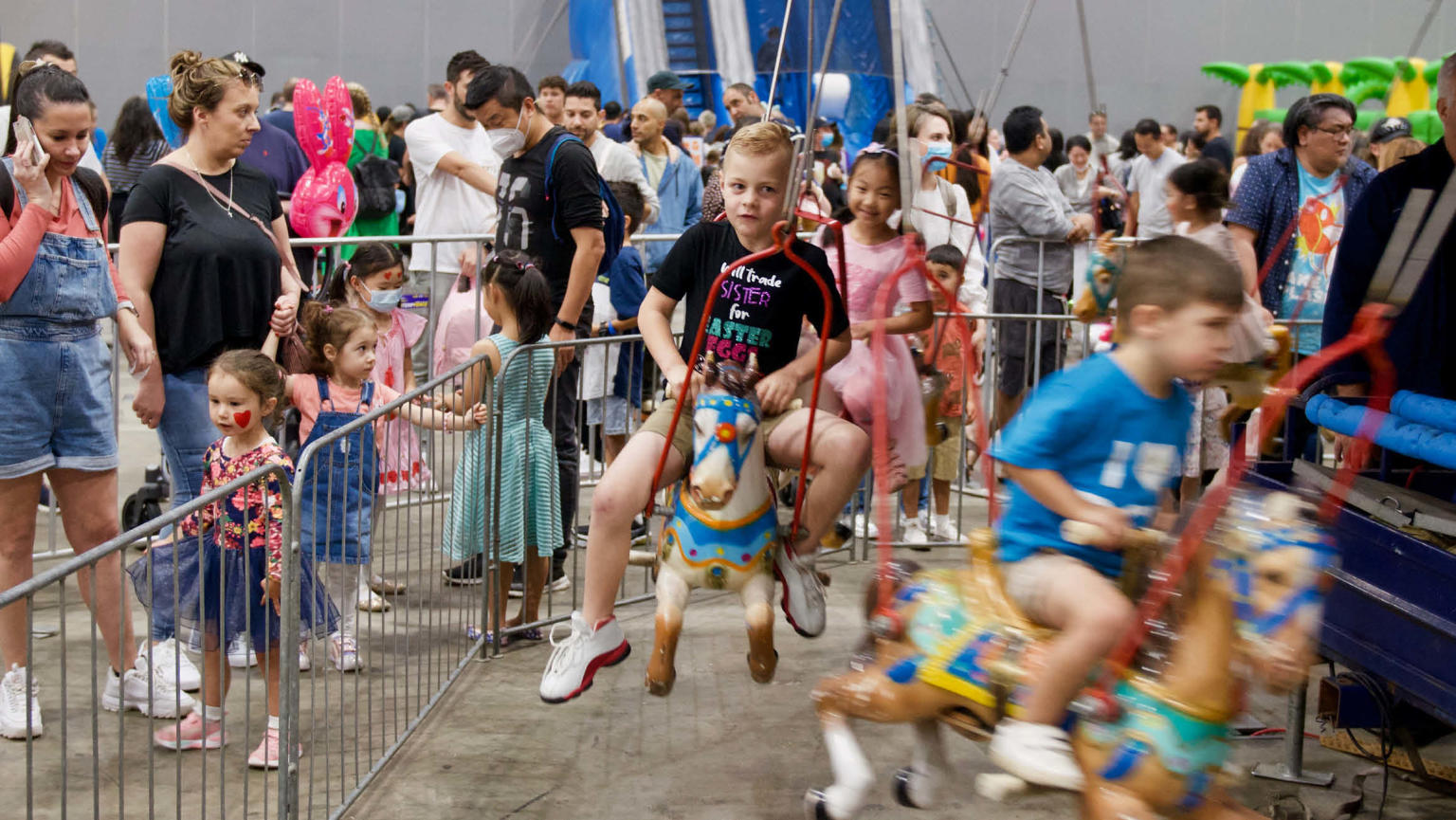 Children sitting on a horse carousel with a busy exhibition of people walking around in the background. 
