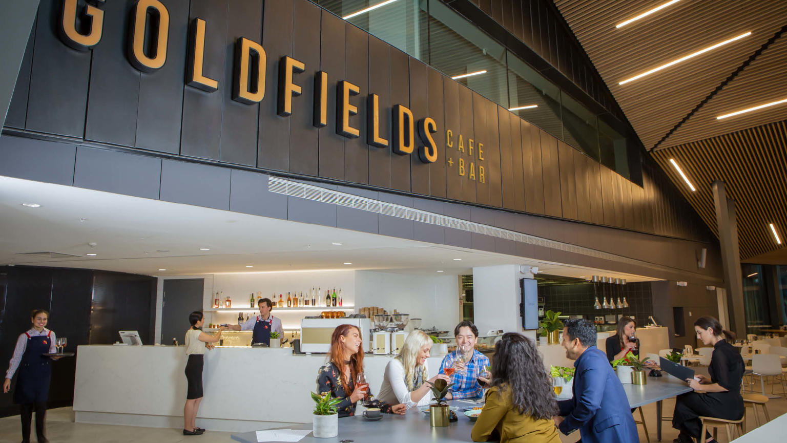 A vibrant café scene, where a group of people enjoys their time seated. Above them, a prominent sign displays 'Goldfields Cafe + Bar'. Behind the bar counter, a barista engages in conversation with a customer while preparing coffee. On the floor, another barista can be seen serving coffee.