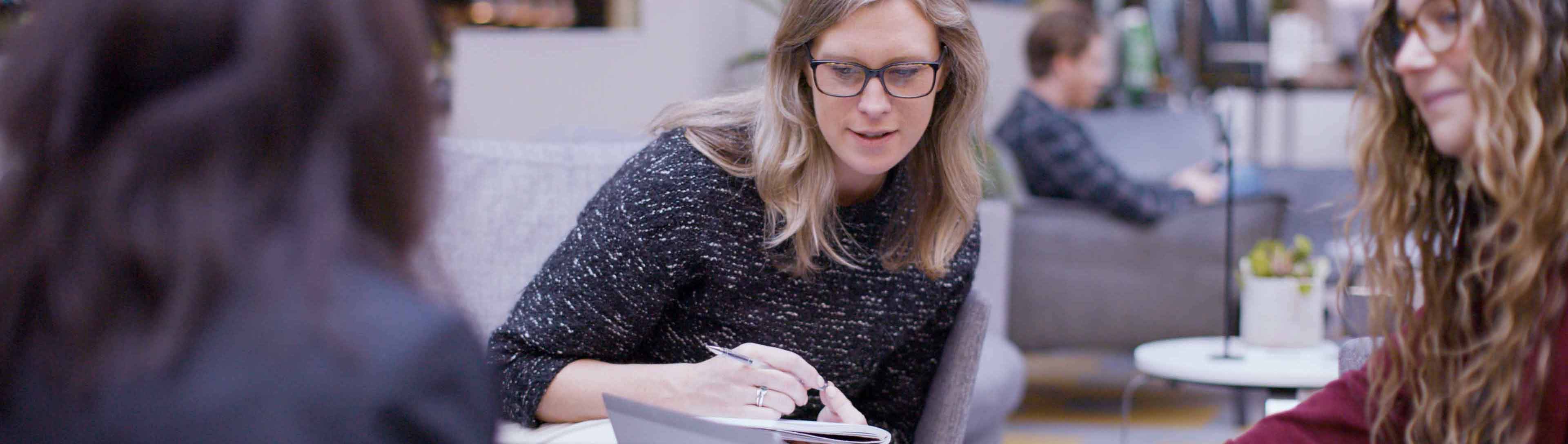 A woman with glasses sits at a chair, she holds a pen in hand looking down at something of camera. In the foreground the shape of two other people can be seen facing the woman in focus. 