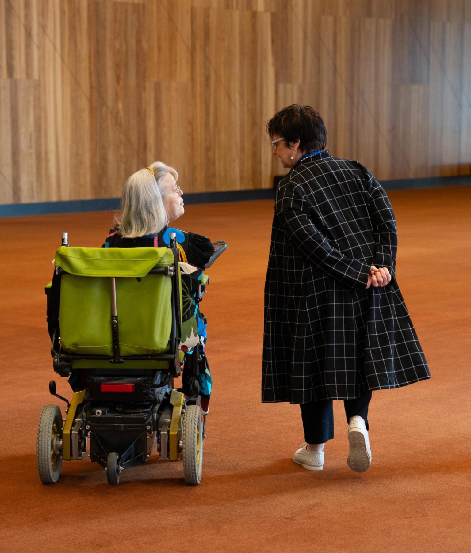 Two woman talking in a hallway with orange carpet. One women walks to the side while the other is in a motorised wheelchair.