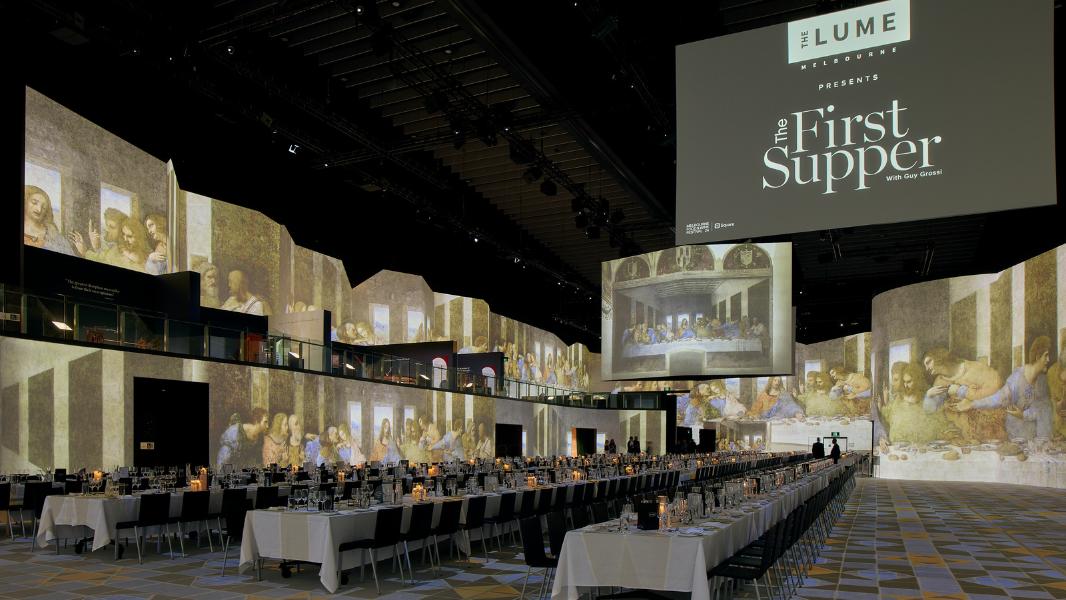 Three long tables running down a large open event space. THE LUME and The First Supper is displayed on a large screen hanging from the roof. 