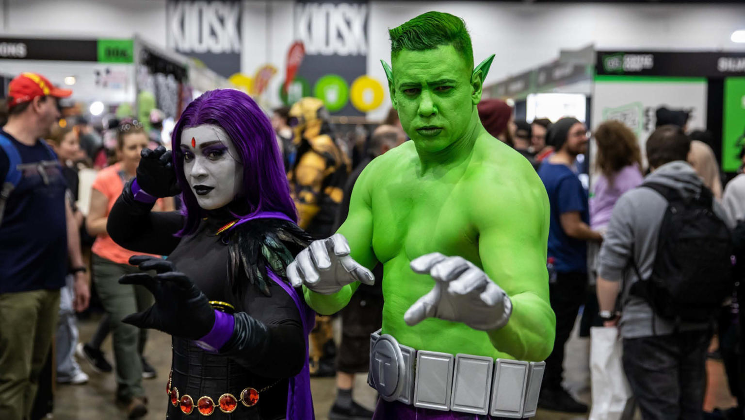 An image showing two people, a man and a woman dressed up at Comic Con. The woman is wearing a purple wig and black costume with a purple cape and red jewelled belt. The man is painted green with elf ears and silver gloves and a belt. 