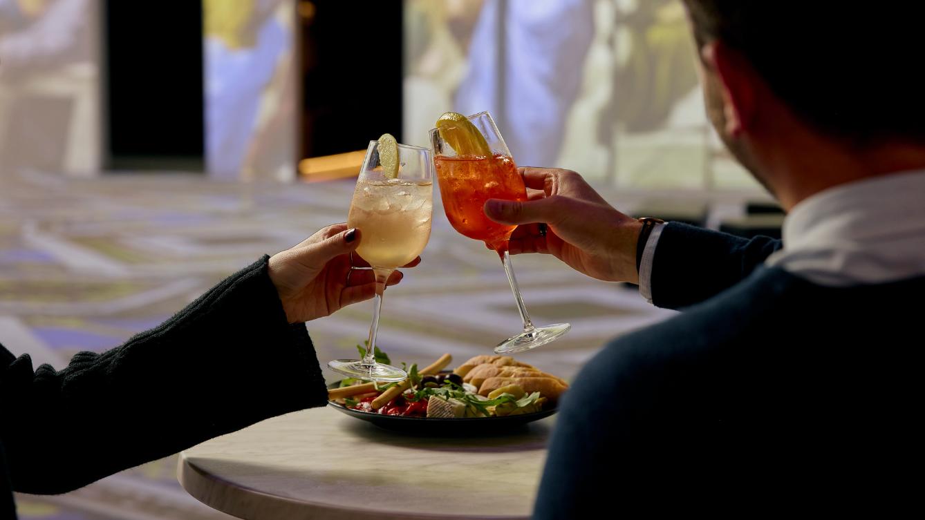 Two people toasting with two cocktails with a dish of food on the table in front of them. 