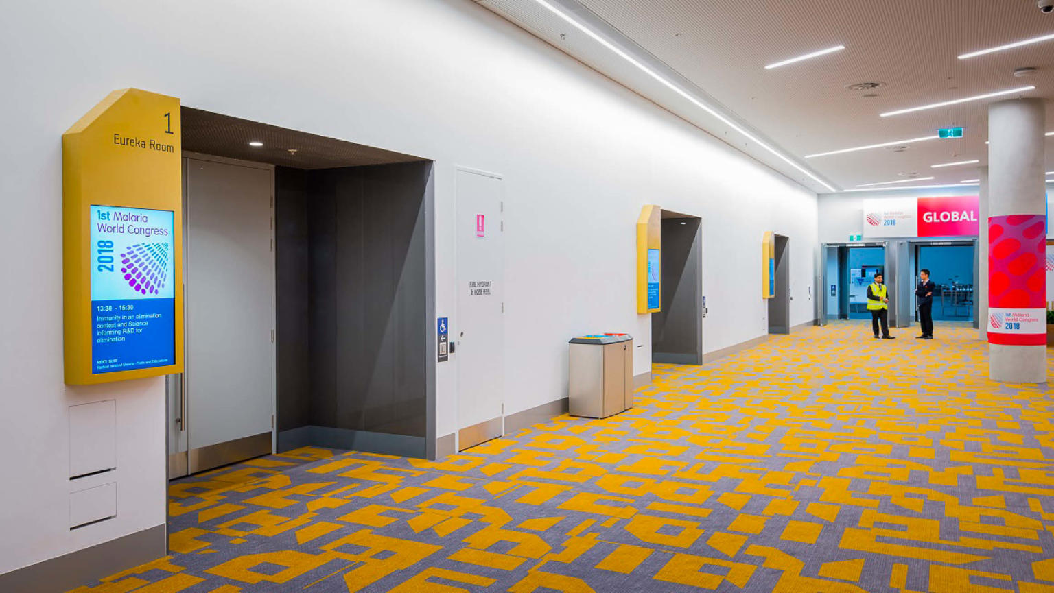 A spacious foyer with multiple room entrances. The carpet displays a combination of yellow and grey tones, while eye-catching bright red and pink signage adorns the surroundings.
