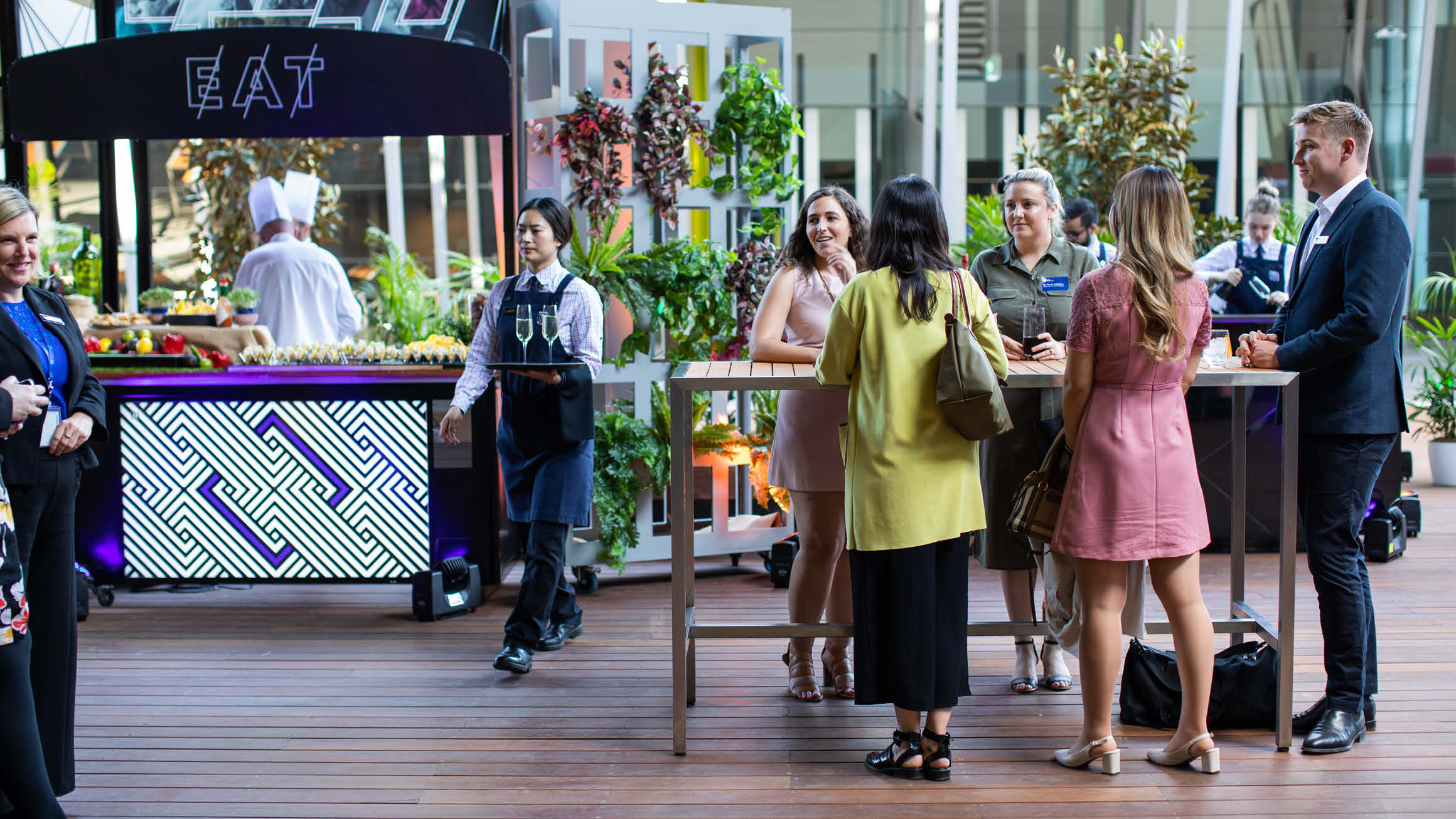 Vibrant courtyard gathering with people socialising around a tall table, enjoying champagne served by a waitress. In the background, a food stall with chefs standing behind it and an 'eat' sign adds to the lively atmosphere.