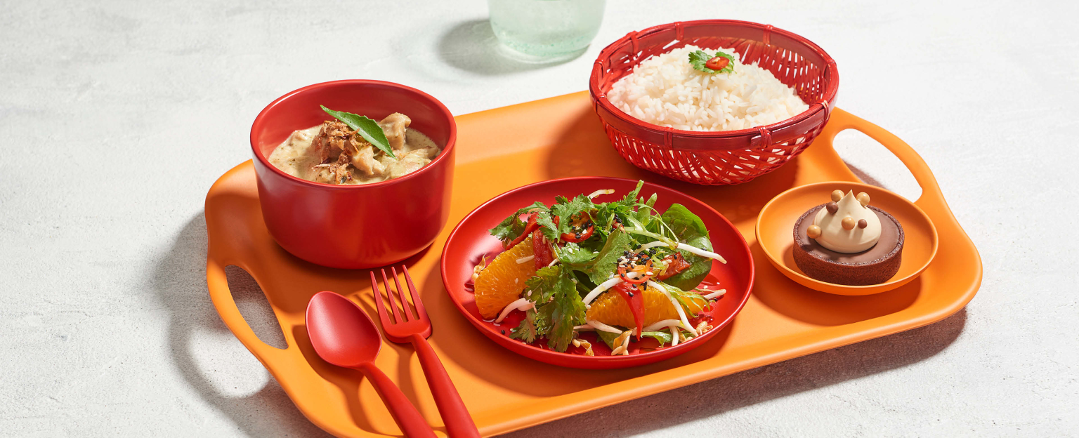 A vibrant orange platter showcasing four dishes: three red bowls, each filled with a unique culinary creation - steamed rice, flavourful curry, refreshing salad, and an orange bowl with a delectable tart. The image is complemented by a set of matching red cutlery.