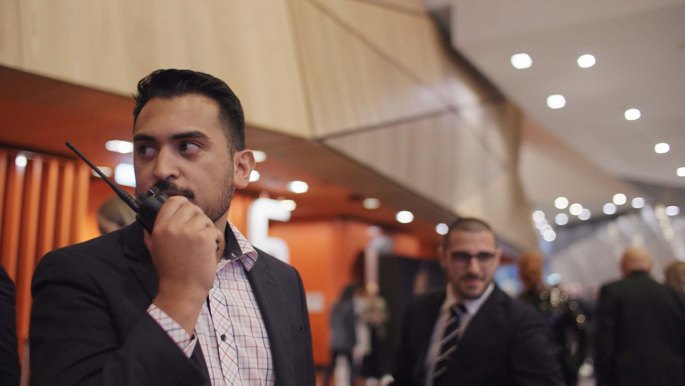 A man wearing a shirt and blazer communicates through a walkie-talkie in a spacious event venue, surrounded by a crowd of people.