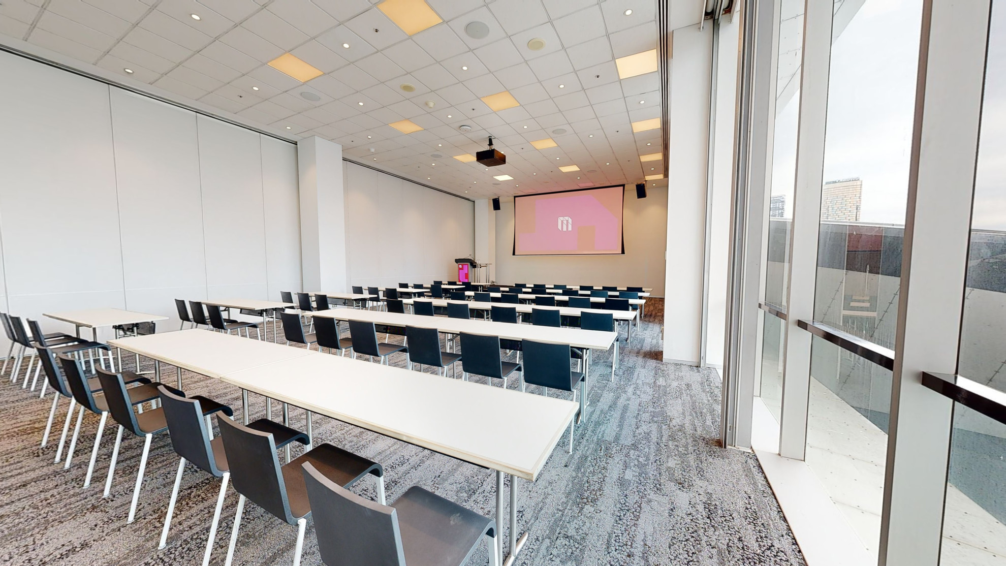 A professional conference or meeting room featuring a spacious layout, equipped with a large screen and comfortable chairs for meetings and presentations. A large floor to ceiling window fills one wall of the room, which looks out to the Melbourne city. 