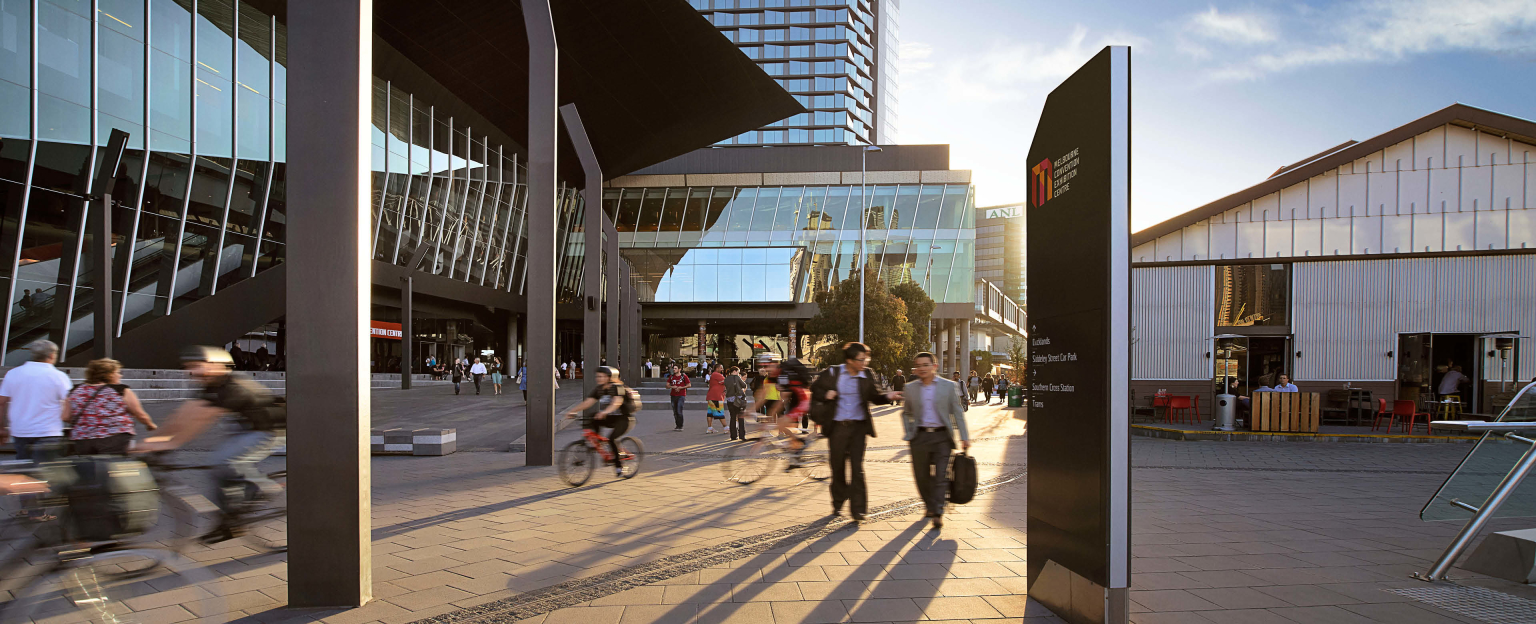 A captivating view of Melbourne Convention and Exhibition Centre from the South Wharf precinct. Bathed in sunlight, the scene showcases people leisurely riding bikes and strolling along the promenade.