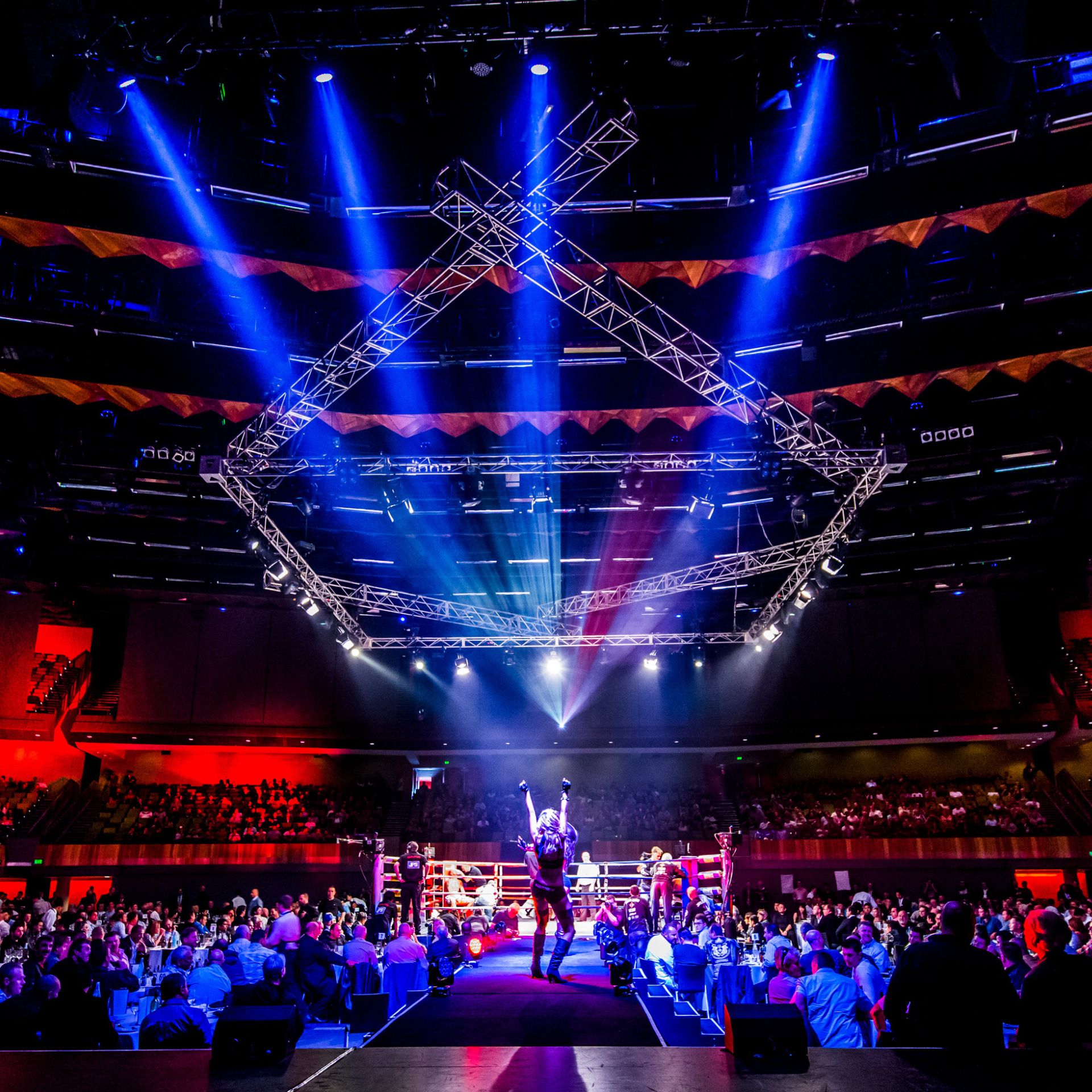 Crowd of people sitting at tables surrounding a boxing ring. Four main spotlights are pointing towards one person standing on a stage with their hands up. 