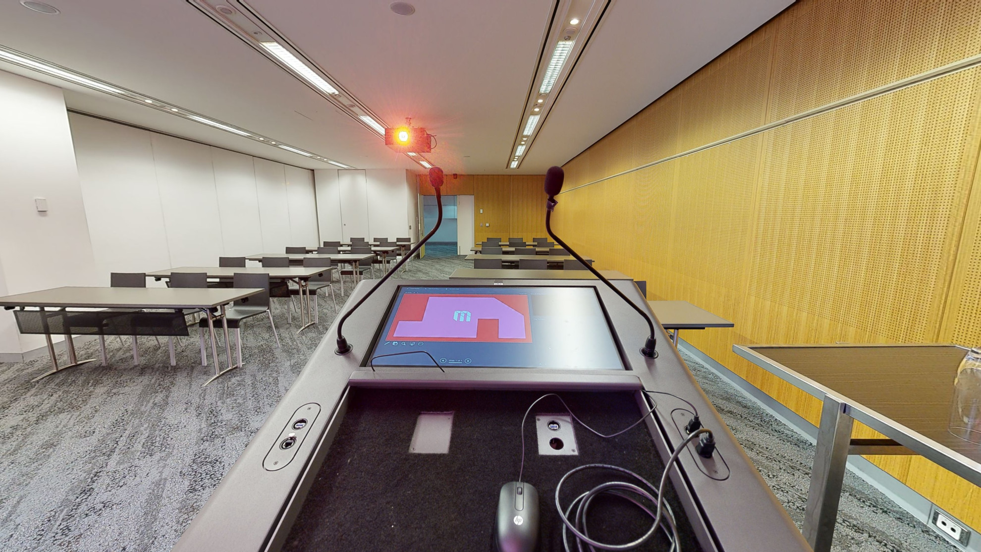 A conference or meeting room featuring a spacious boardroom layout equipped with a sizable screen and comfortable chairs for attendees. A lectern also sits in the corner to the left of the screen next to a large floor to ceiling window. 