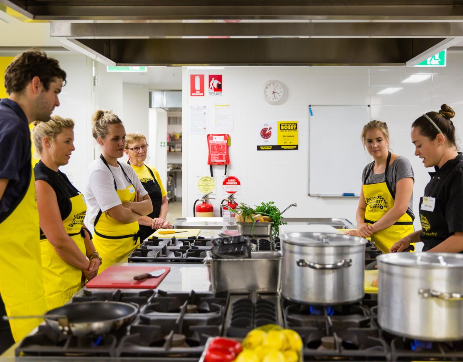OzHarvest cooking class - A group of people gather around a kitchen bench and stove, listening to a chef gives directions on preparing the food. 
