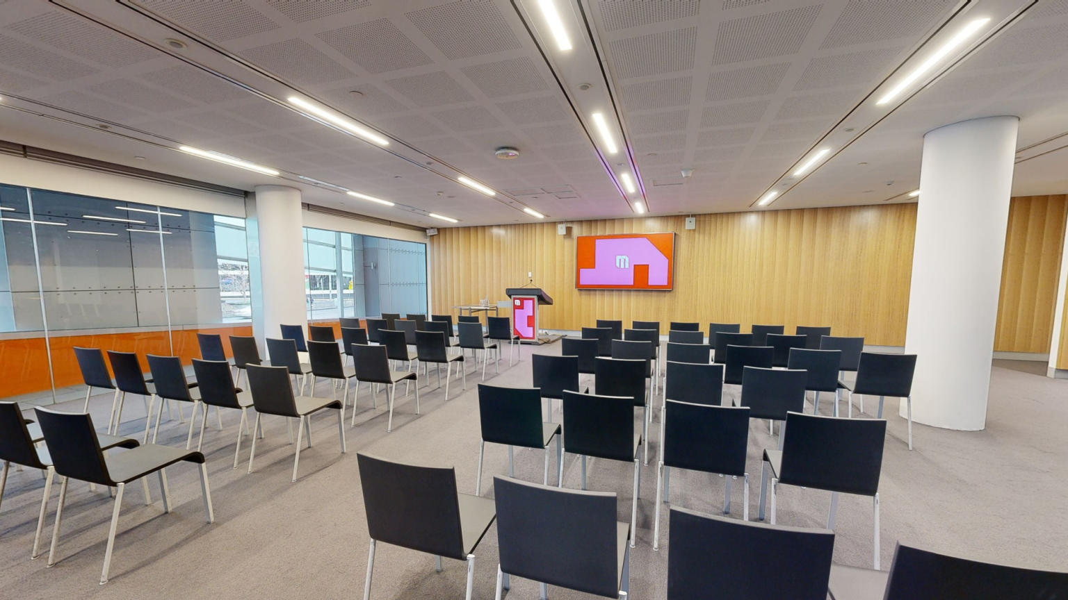 A conference or meeting room featuring rows of chairs arranged to face large tv screen for presentations and discussions. A lectern sits to the side of the tv screen and windows run along the side of the room facing out towards Crown. 