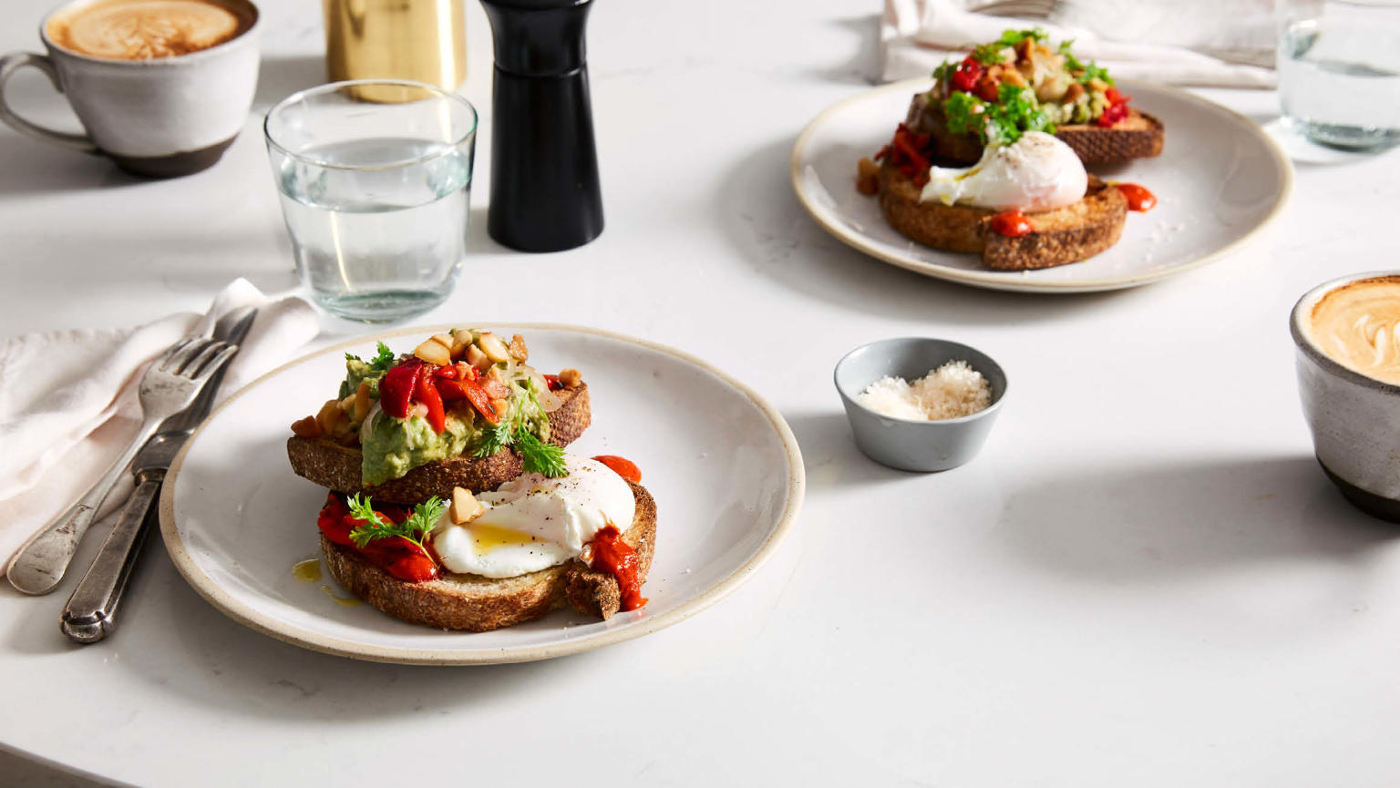 An image featuring a beautifully presented breakfast spread on a table. There are two plates with toast, creamy smashed avocado, poached eggs, and a roasted capsicum garnish. The table is further adorned with cups of coffee, neatly arranged cutlery, salt and pepper shakers, and a small bowl of grated cheese. 
