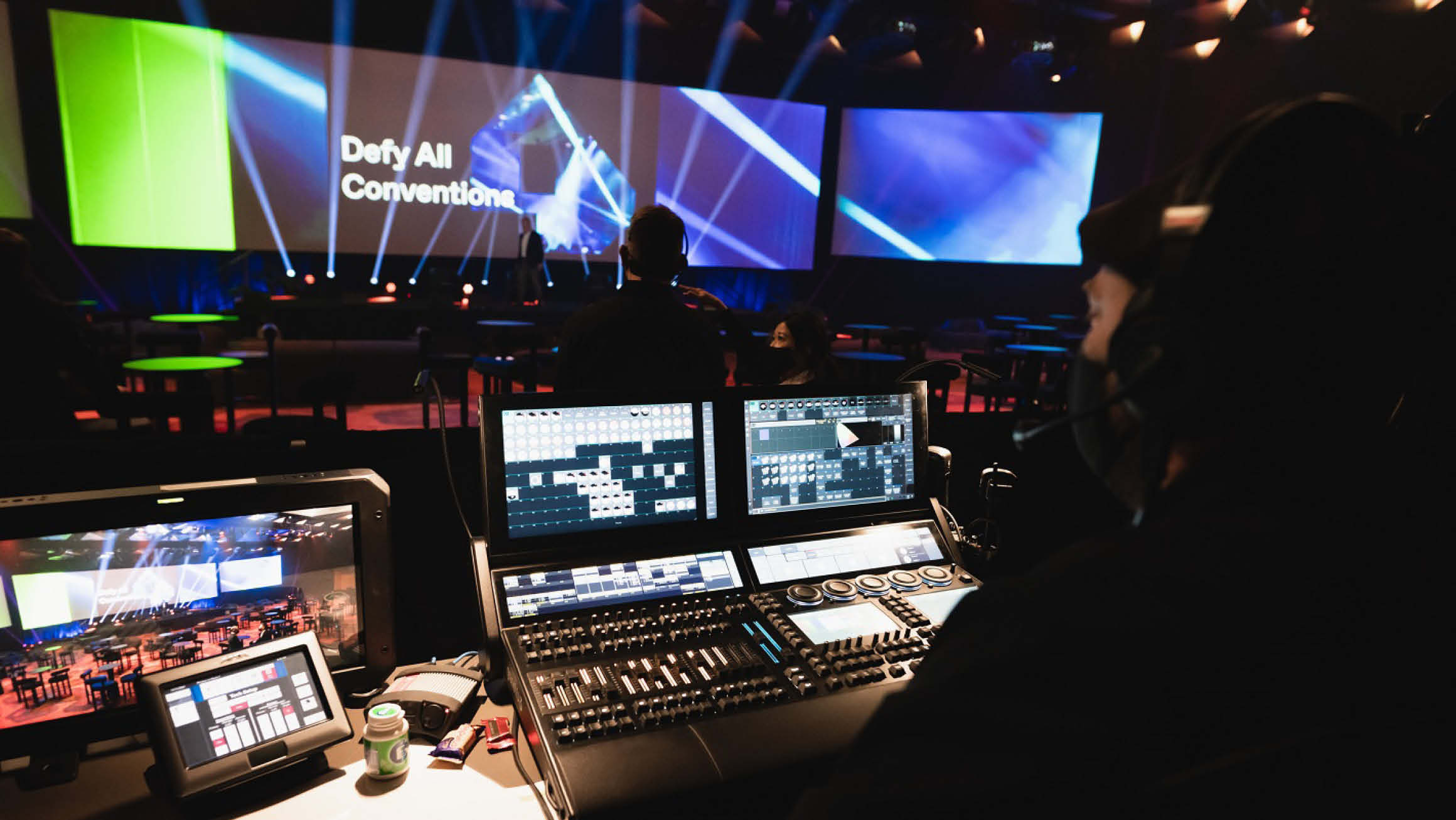 Looking over a person's shoulder who sits in the shadow of a dark room. They look upon a busy technology board with lots of screens along the wall. sitting at eye level. 