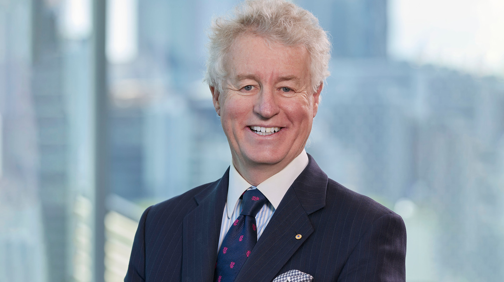 A close up headshot of Peter Jones - a man with white hair smiling wearing a black suit with a blue tie and white collared shirt. 