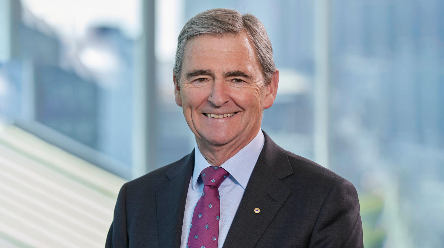 A close up of John Brumby - a grey haired man wearing a black suit over a pale blue collared shirt with a purple tie.