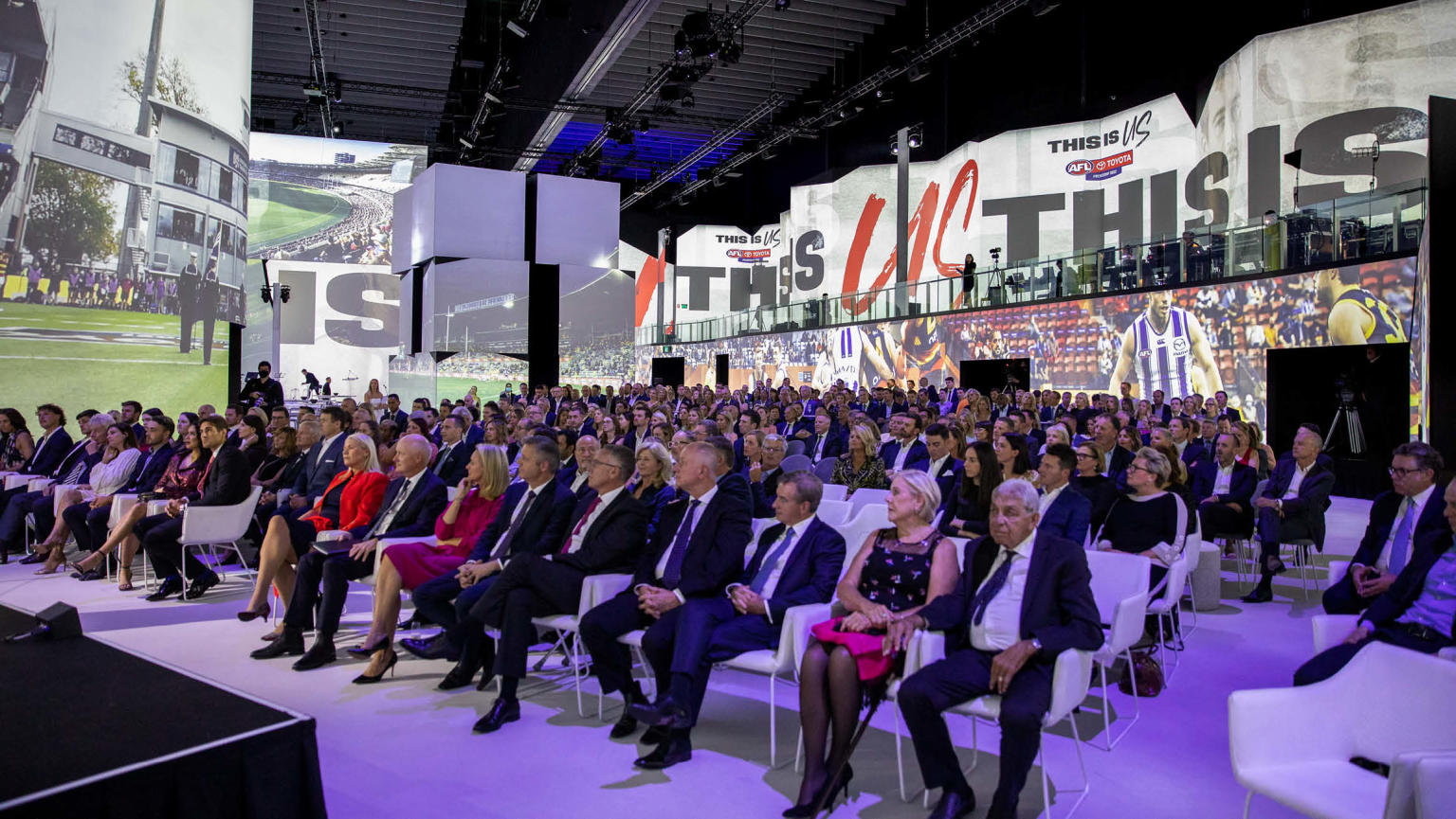 A seated audience in rows, attentively facing a stage, while immersive digital screens adorn the surrounding walls.