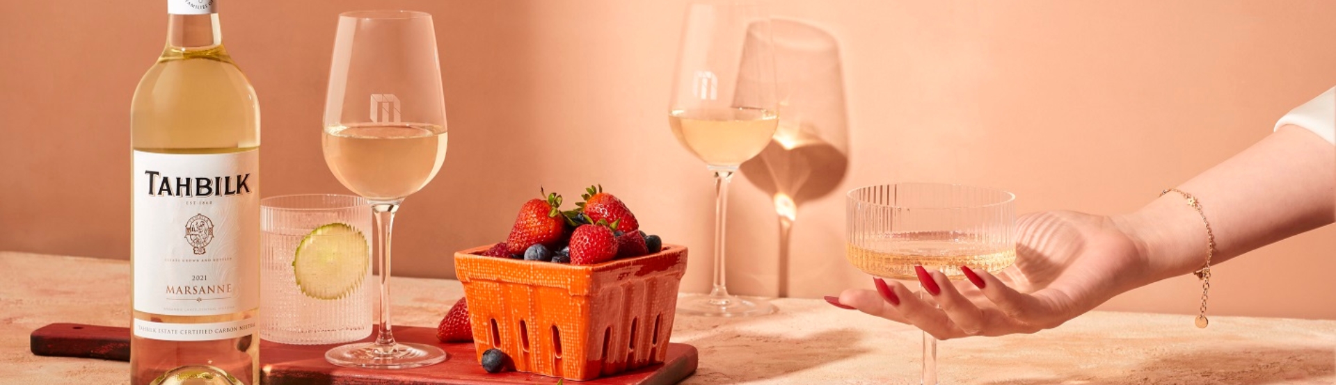 A Tahbilk bottle of white wine next to a collection of wine and cocktail glasses filled is scattered on the table with an orange carton of fresh berries. 