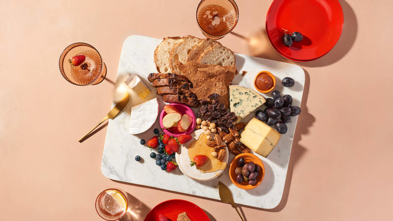A appetising display of assorted breads, cheeses, nuts, and fruits elegantly arranged on a marble platter atop a peach table. The image also includes two vibrant red plates, two champagne coupes, a tall glass and gold cutlery.