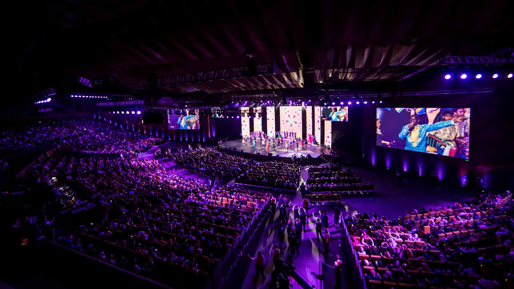 A large theater filled with people. The view is from the back top corner of the theater, looking down onto the stage. The stage is brightly lit, and there is a performance happening on it. The audience is sitting in the seats, and they are all paying attention to the performance.