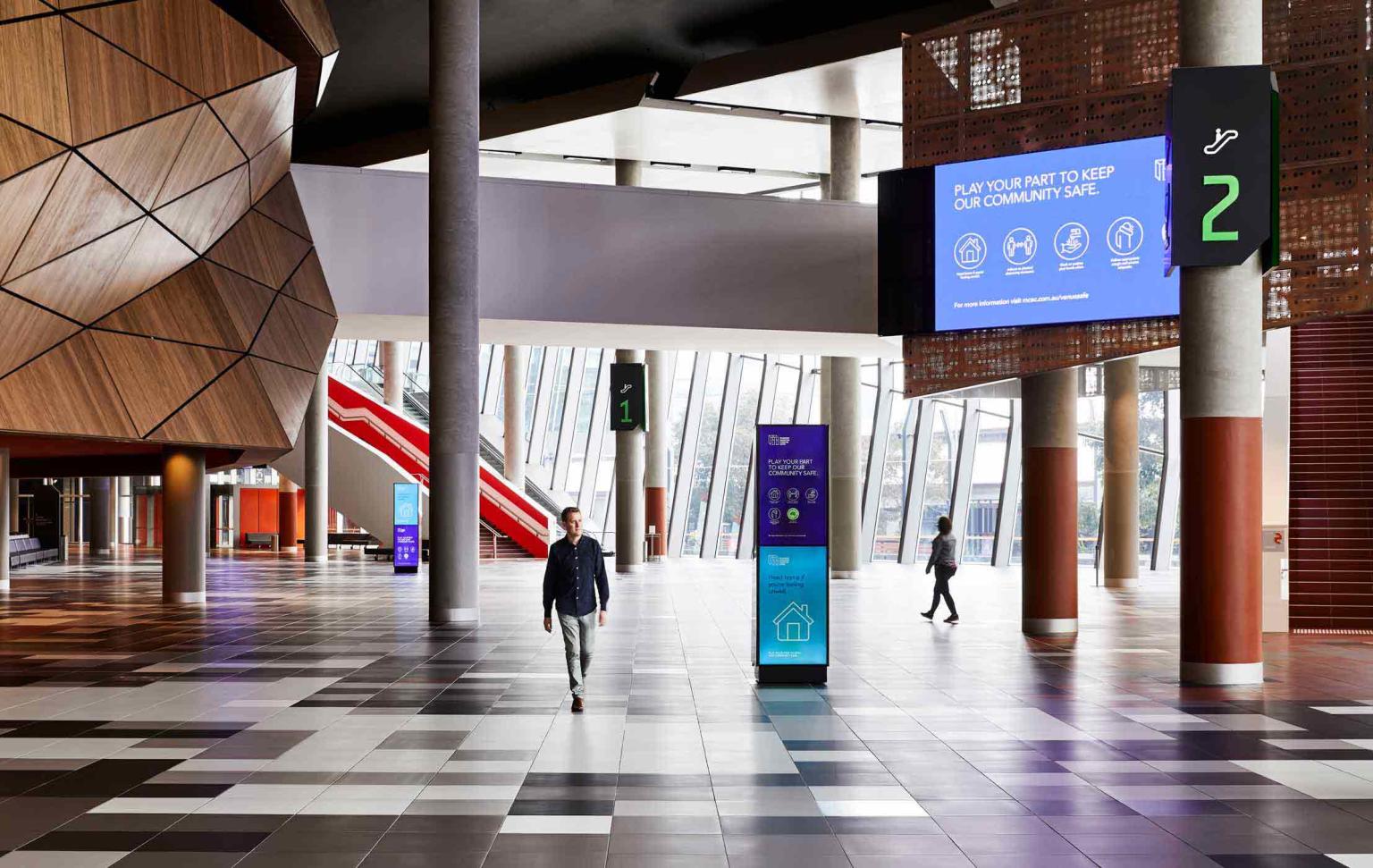 A man is walking through the empty foyer of the Melbourne Convention Centre. A large screen in the background displays covid safe directions. 