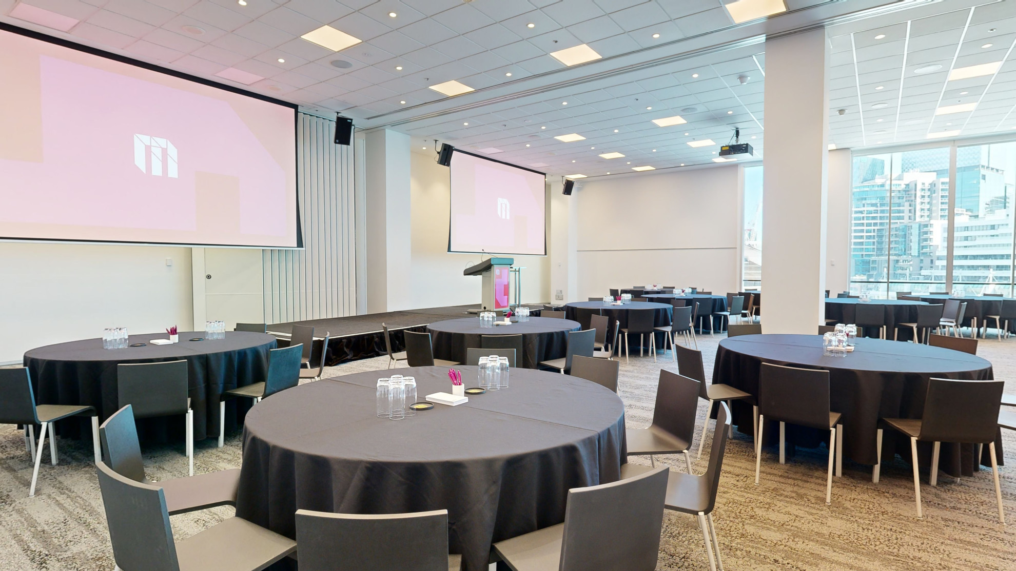 A spacious conference room with tables and chairs arranged for a meeting facing two large screens at the front of the room. Large floor to ceiling windows are to one side of the room. 