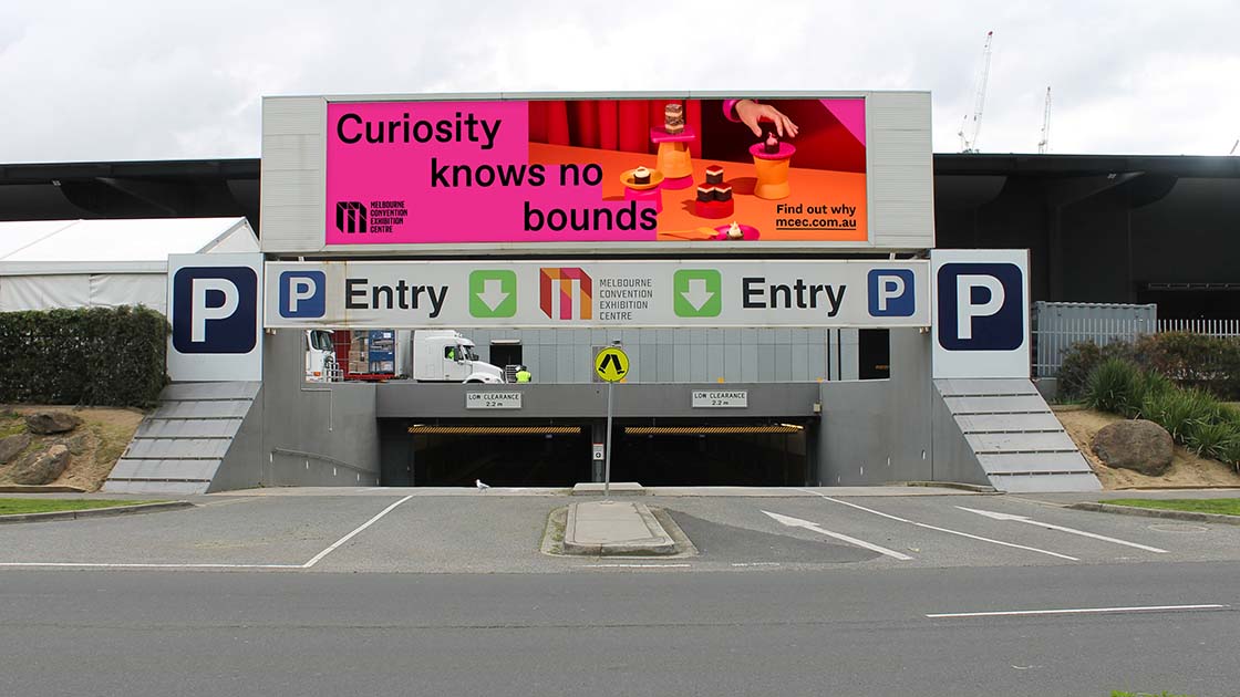 A concrete parking lot entrance marked by a prominent green sign overhead. The sign bears the humorous phrase "Nothing like 5,000 strangers to make you feel at home," accompanied by the Melbourne Convention Exhibition Centre logo in the bottom left corner.