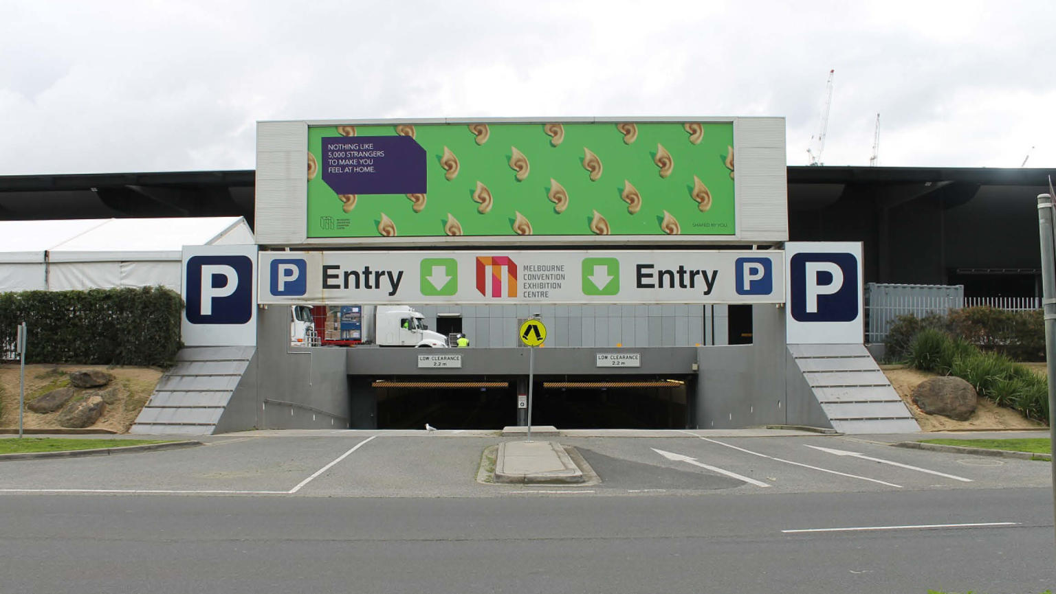 A concrete parking lot entrance marked by a prominent green sign overhead. The sign bears the humorous phrase "Nothing like 5,000 strangers to make you feel at home," accompanied by the Melbourne Convention Exhibition Centre logo in the bottom left corner.