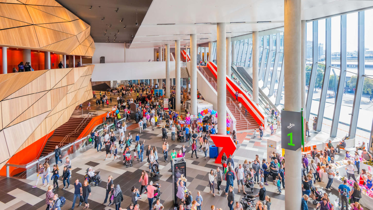 Image of a large foyer area within the Melbourne Convention and Exhibition Centre (MCEC). The foyer is filled with people walking around. There are concrete pillars, stairs, large windows and a wooden wall. The foyer is well-lit and appears to be a busy place.
