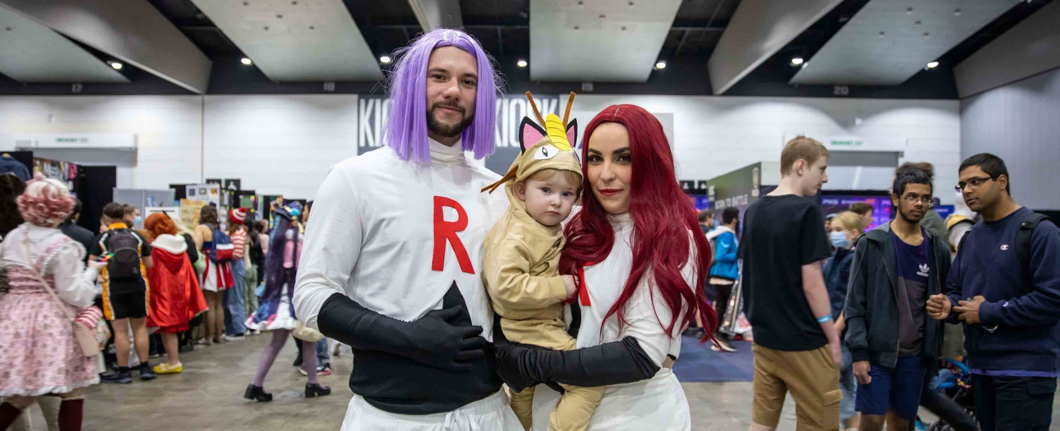 This image shows a family dressed up in costumes at Comic Con. The man is wearing a white costume with a purple wig, the woman is wearing a white costume with a deep red wig, and the child is wearing a light brown costume. They man and woman are also wearing long black gloves. The people in the background are also dressed up in costumes and are walking around the exhibition bays.