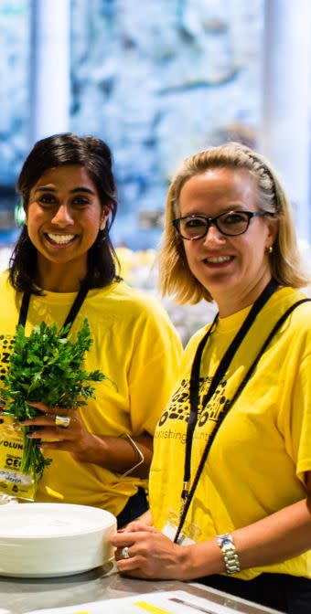 Two women in yellow Ozharvest t-shirts, one holding a bunch of herbs, smiling at the camera. 
