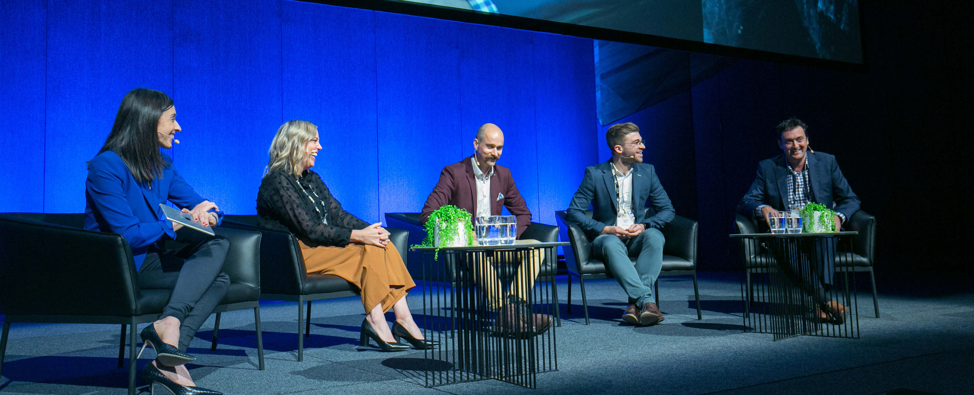 A panel discussion on a stage featuring five individuals seated in armchairs. The participants engage in active conversation, exchanging thoughtful glances as they interact with one another. 