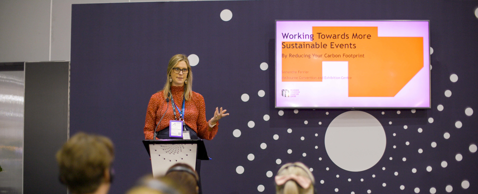 Sam Ferrier, wearing an orange jumper, presenting at a podium to a crowd off camera with a screen behind her with the heading displayed "Working towards more sustainable events".